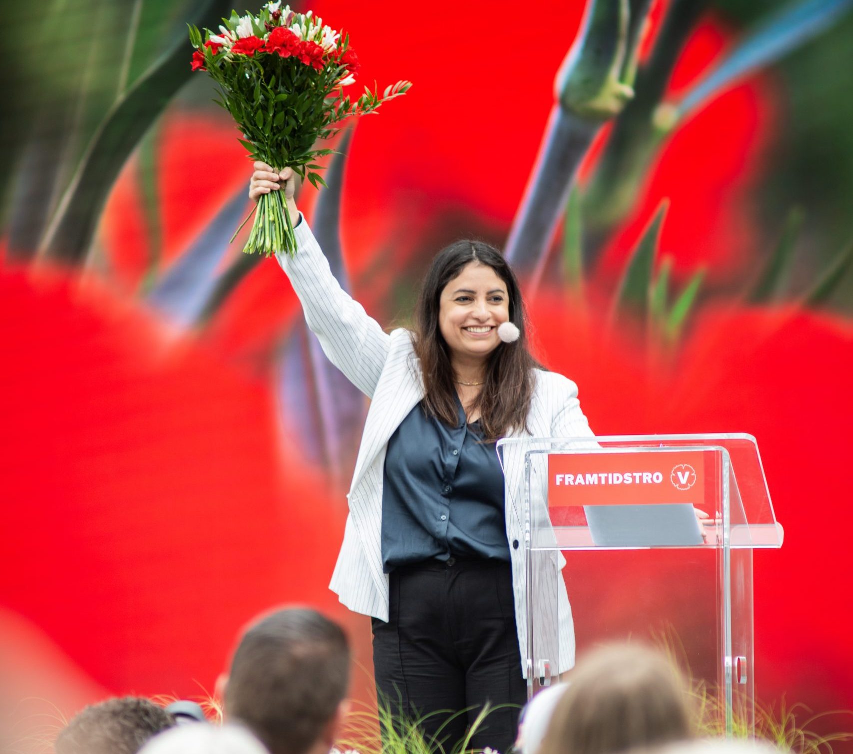 Nooshi Dadgostar står på en scen bakom ett genomskinligt talarstativ med Vänsterpartiets logotyp. Hon ler och håller upp en blombukett med röda blommor i sin högra hand. Bakgrunden domineras av en stor, konstnärlig bild på röda blommor.