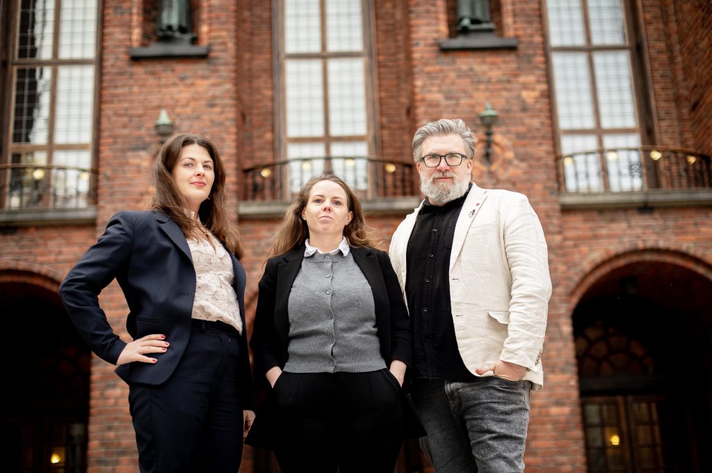 Tre personer V-politiker poserar för en bild framför Stockholms stadshus tegelfasad, med fönster och statyer i bakgrunden. Från vänster: Deniz Butros i mörkblå blazer och spetstopp, Alexandra Mattsson i mitten med grå kofta, svart kavaj och ena vänster hand lyft, samt Robert Mjörnberg till höger i vit linneblazer och glasögon med rak hållning.