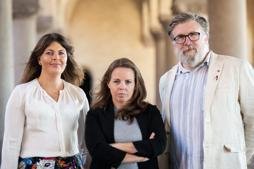 Vänsterpartiets kandidater står utomhus i stadshusets pelargång. Från vänster: Deniz Butros i vit överdel och färgglad, mönstrad kjol med handen på höften, alexandra mattsson i svart kavaj och ljusgrå tröja, samt robert mjörnberg i glasögon klädd i en ljus kavaj över en ljusblå skjorta.