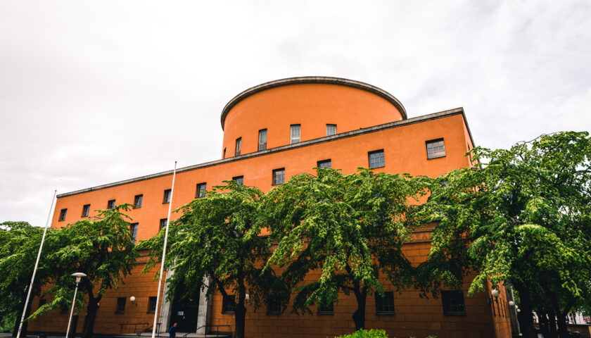 Sommarbild med grönskande träd framför Stockholms stadsbiblioteks oranga fasad.